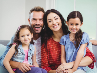 Portrait,Of,Cheerful,Parents,Sitting,With,Daughters,On,Sofa,At Family Portrait